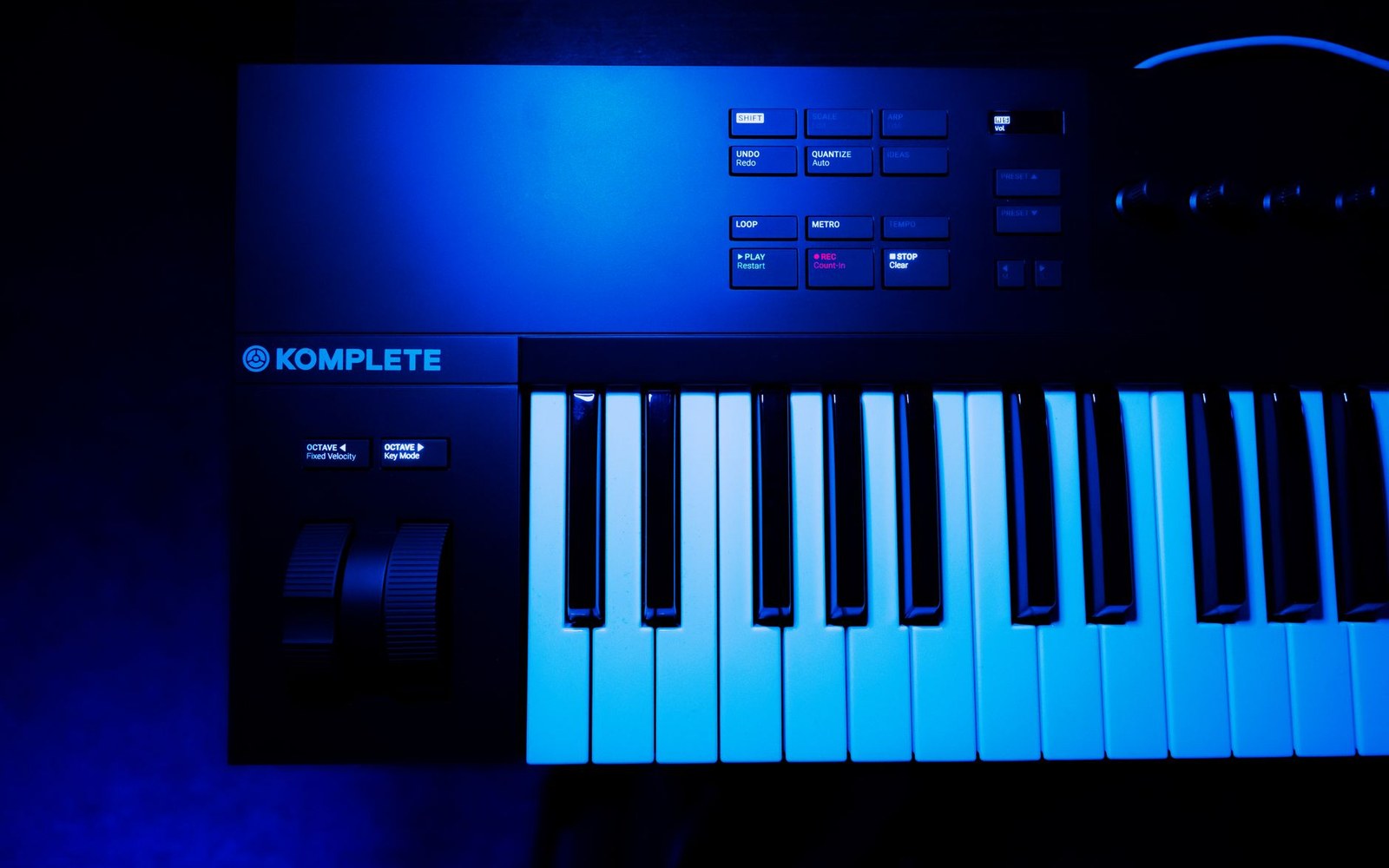 Blue-lit Komplete keyboard controller showing transport buttons and piano keys close up
