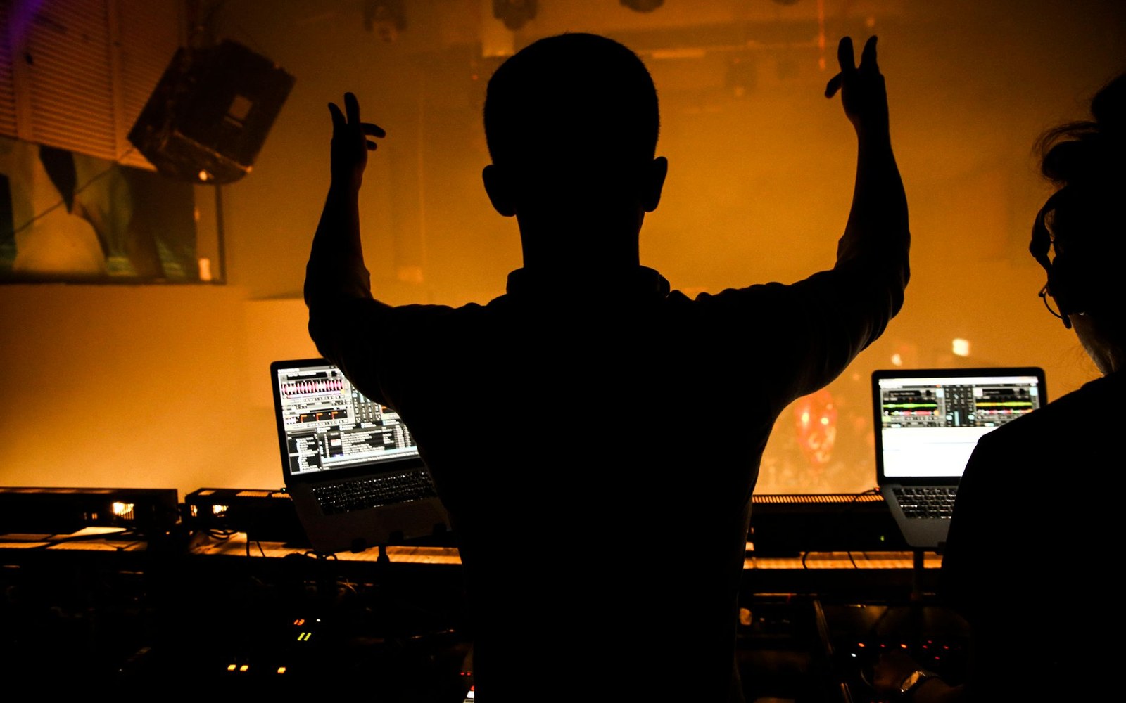 Silhouetted DJ raises both hands amid orange stage haze, laptops glowing on the decks