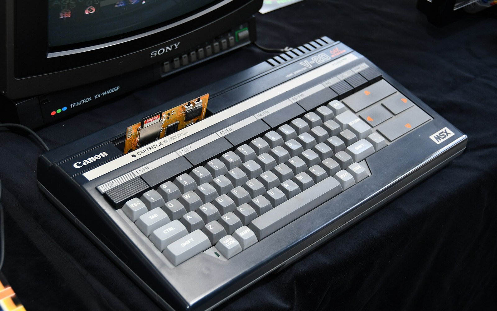 A Canon MSX computer keyboard with an exposed cartridge circuit board, beside a Sony Trinitron monitor